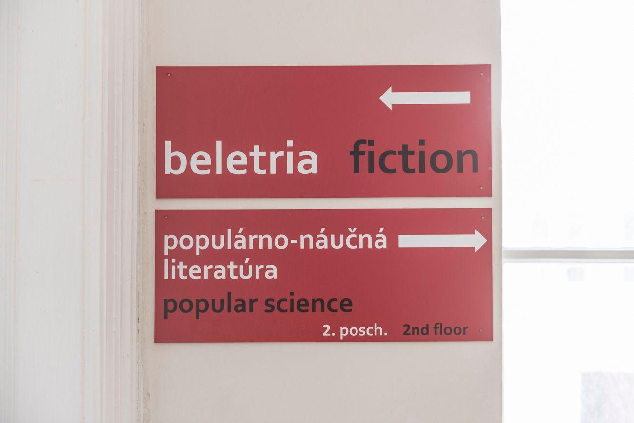 Popis fotografie: Na stene je zavesená tabuľa s popisom a šípkami smeru rozdelenia kníh na beletria-fiction (šípka smer vľavo) a populárno-náčná literatúra-popular science (šípka smer vpravo) 2. poschodie-2nd floor.