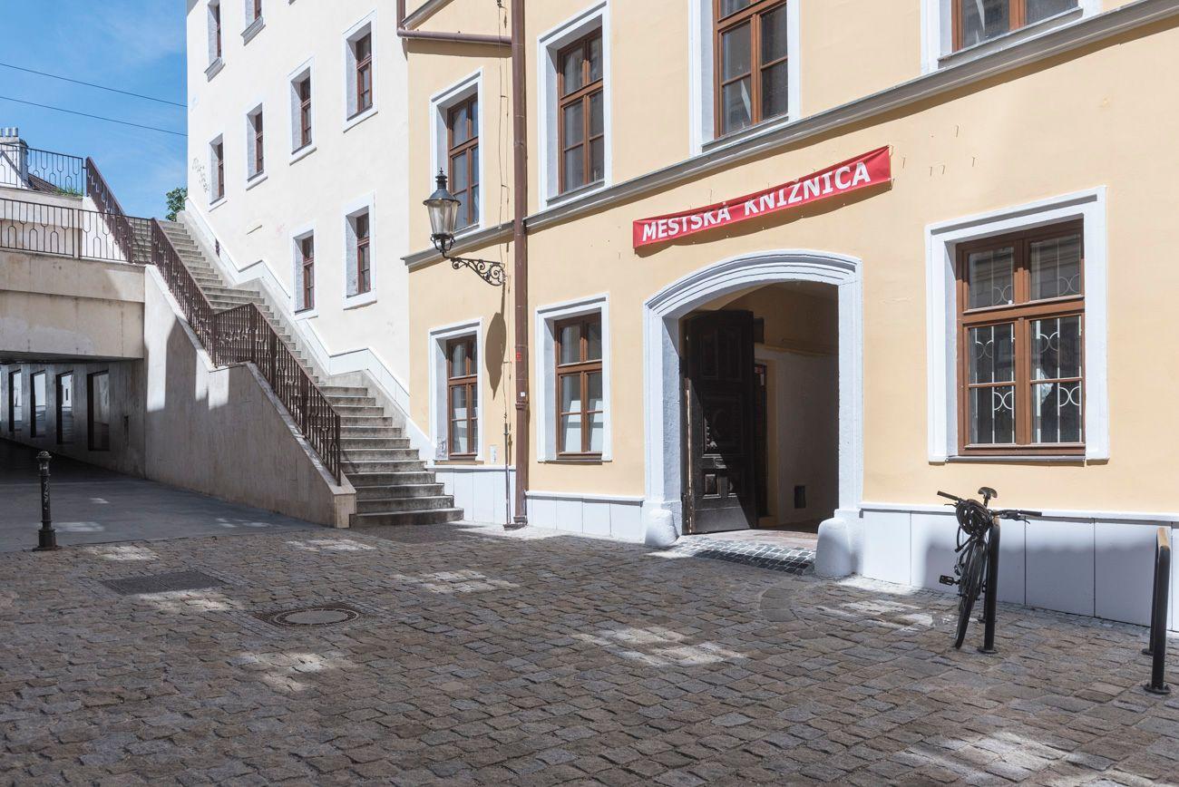 Popis fotografie: Na obrázku je zobrazený vchod z ulice do budovy. Nad vstupnou bránou je nápis Mestská knižnica. Pred budovou je opretý bicykel o stojan na bicykle.