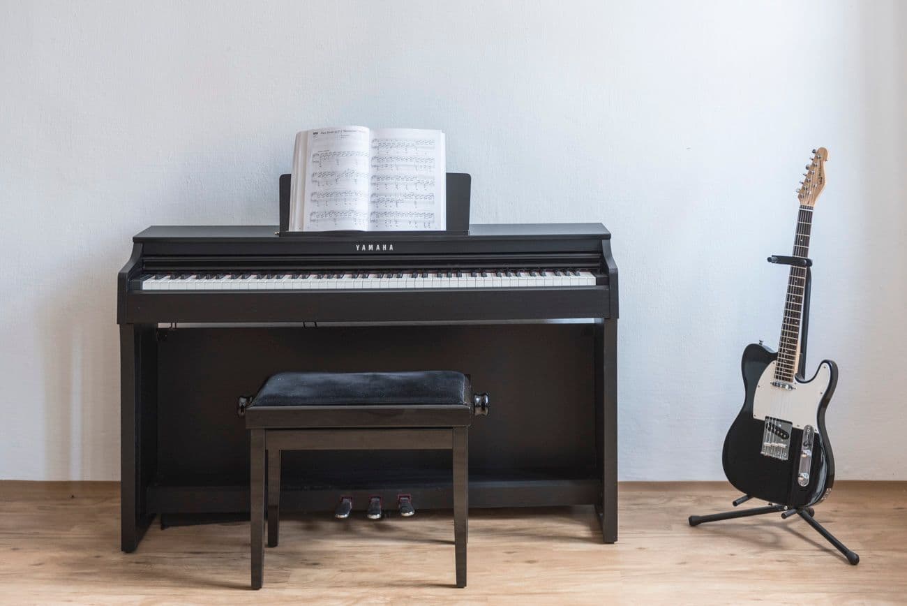 Popis fotografie: Záber na klavír so stoličkou doplnený o hudobné noty. Vedľa klavíra je v stojane odložená elektrická gitara.